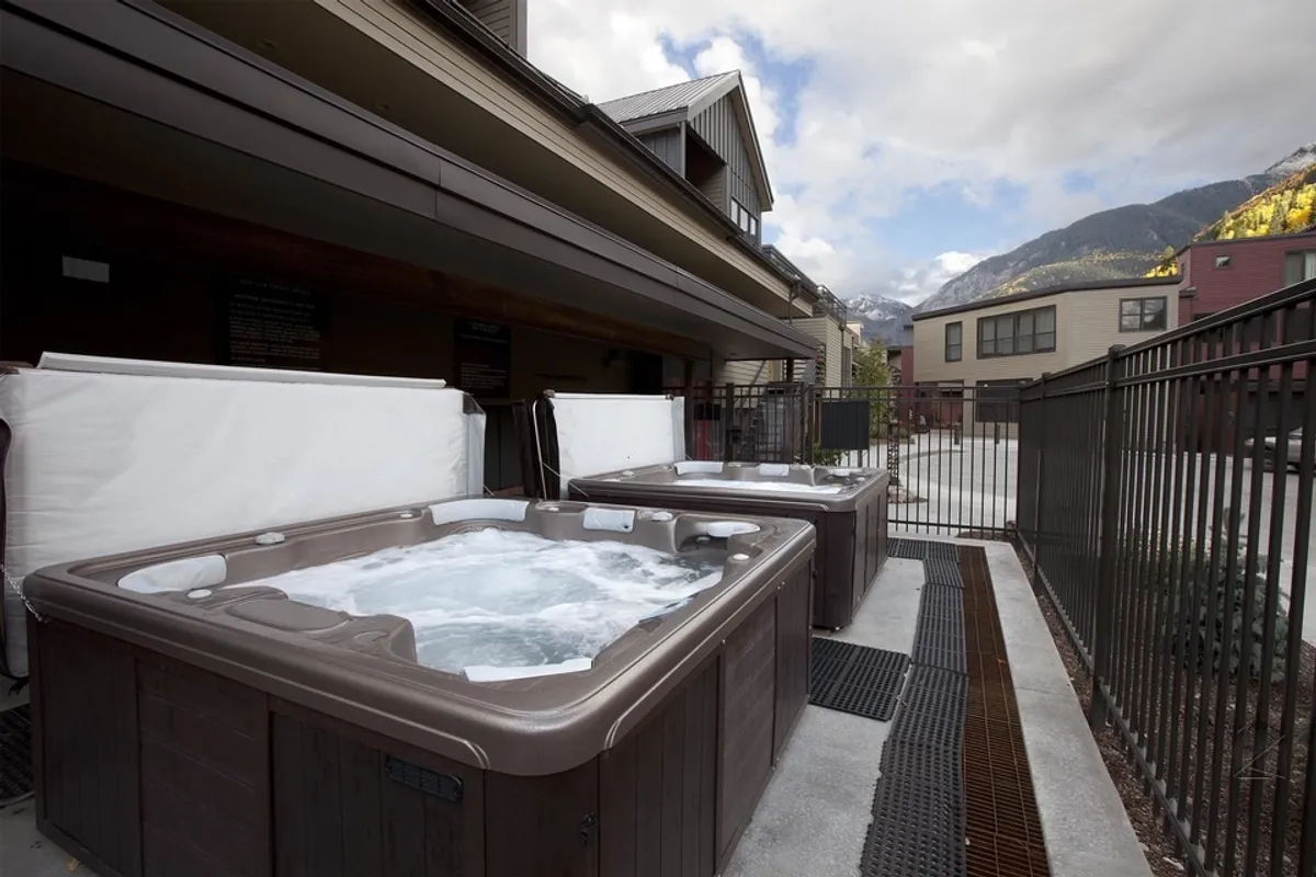 This image shows two outdoor hot tubs nestled against a picturesque mountain backdrop, with surrounding lodge buildings in the frame. The setting is tranquil, emphasizing relaxation and the resort-like atmosphere of the property. The hot tubs are HOA-managed amenities described in the section, and the mountain views reinforce the Telluride experience.