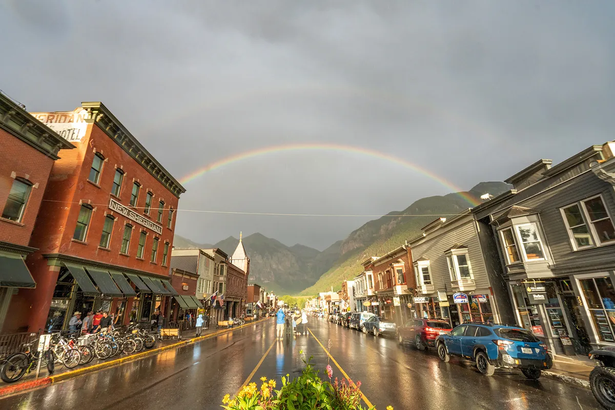 A vibrant Telluride street scene after rain, with historic buildings, a visible rainbow, wet streets, and people strolling. The setting conveys both access to local shops and a community atmosphere—ideal for family outings to nearby attractions like Town Park and Clark’s Market.