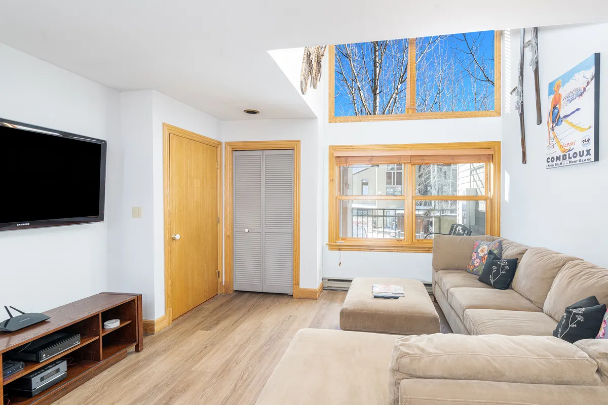 A cozy living room with a comfortable sofa positioned to face a large window, which frames sweeping mountain views. A flat-screen television is mounted nearby, and the room is brightened by ample natural sunlight. The setting encourages relaxation and conversation, making it ideal for après-ski gatherings or a family movie night.