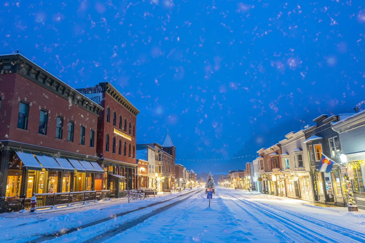 A charming snow-covered street in Telluride’s historic downtown is illuminated by shop windows and a sparkling Christmas tree, with pedestrians enjoying a magical winter atmosphere. The inviting storefronts and festive lighting highlight the ease of reaching Main Street and local eateries by foot.