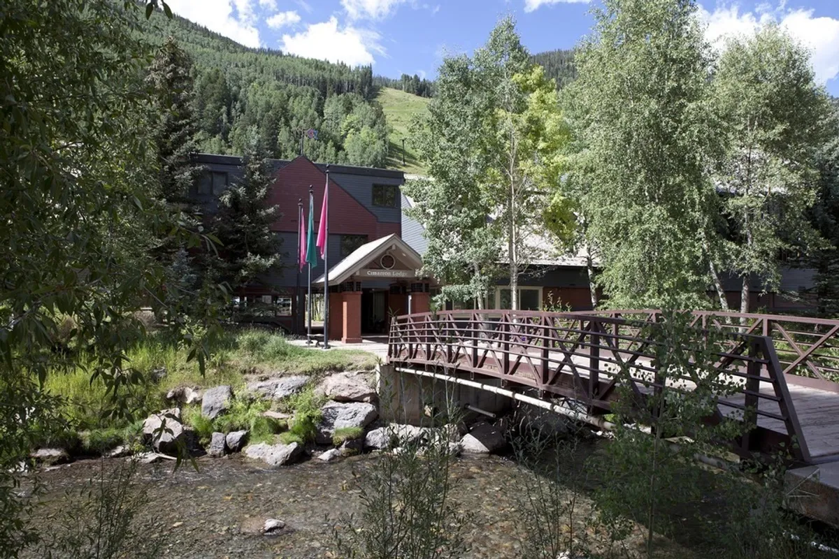 A picturesque wooden bridge spans a flowing stream just outside the building's facade. Verdant greenery surrounds the area, inviting families to enjoy nature walks, observe wildlife, and relax by the gentle water. The building in the background hints at close proximity to the lodge.
