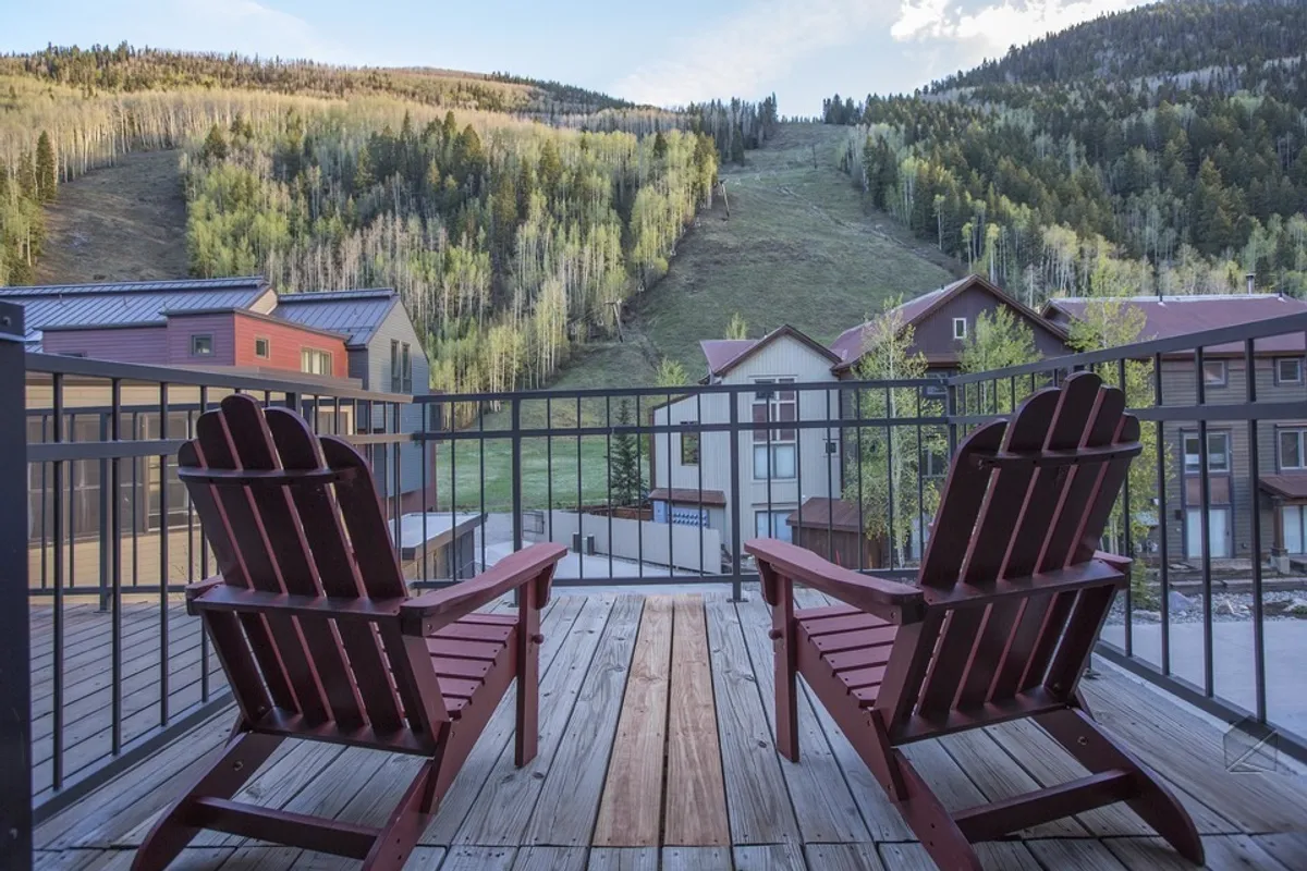 The image features two inviting red Adirondack chairs positioned on a deck, overlooking a breathtaking mountain landscape. The modern condo buildings of Cimarron Lodge are visible, setting the scene for a relaxing retreat. The overall mood is serene, with the setting sun casting a warm glow and suggesting possibilities for sunset views, stargazing, or cozy outdoor gatherings.