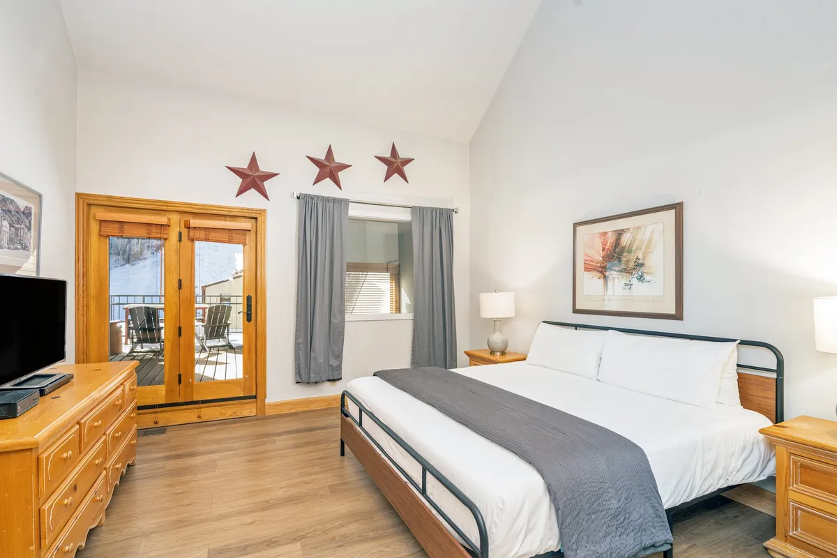A cozy bedroom featuring a plush bed adorned with crisp linens, large windows that let in abundant natural light, a dresser, wall art, and twinkling star accents on the wall. The view from the window hints at the balcony and mountain scenery outside, creating a serene retreat ideal for relaxation.
