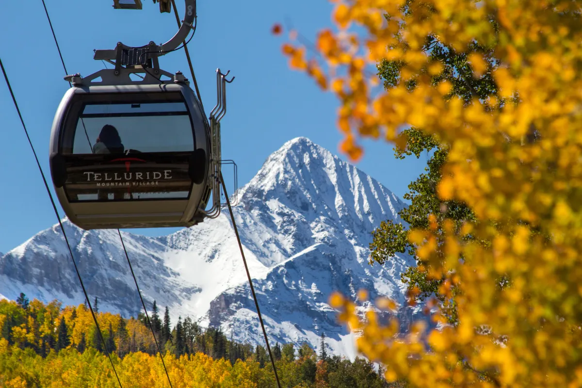 A gondola glides above a snow-capped mountain, framed by vibrant autumn foliage, capturing the beauty of Telluride’s changing seasons and its iconic cable car system. The scene showcases easy access to trails and ski slopes, with nature at your doorstep.