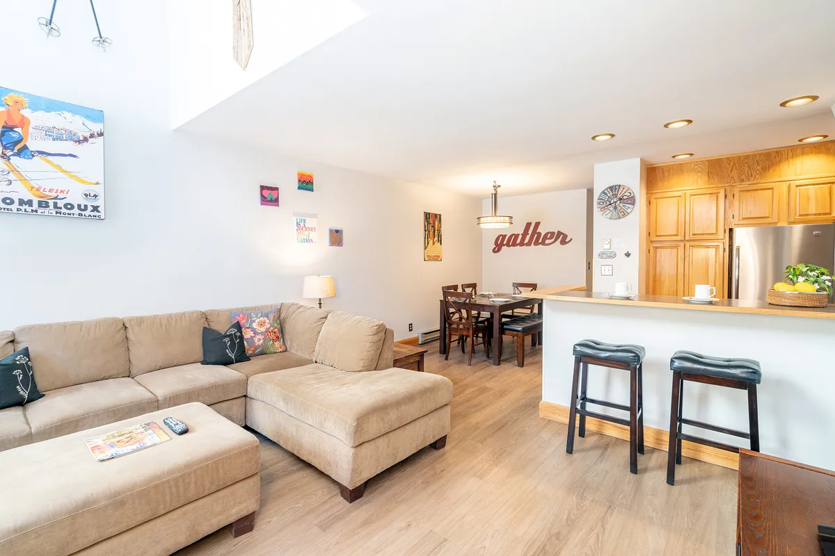 A warm, inviting living space with a plush sofa, stylish dining table, modern counter area with bar stools, and decorative wall art. The room feels comfortable and ideal for relaxation or casual family time.