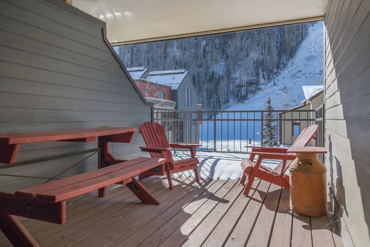 An inviting deck scene with red Adirondack chairs and a wooden bench overlooking snow-covered ground and distant mountains. The setting feels restful, perfect for unwinding after a day on the slopes, with panoramic mountain views enhancing the après-ski atmosphere.