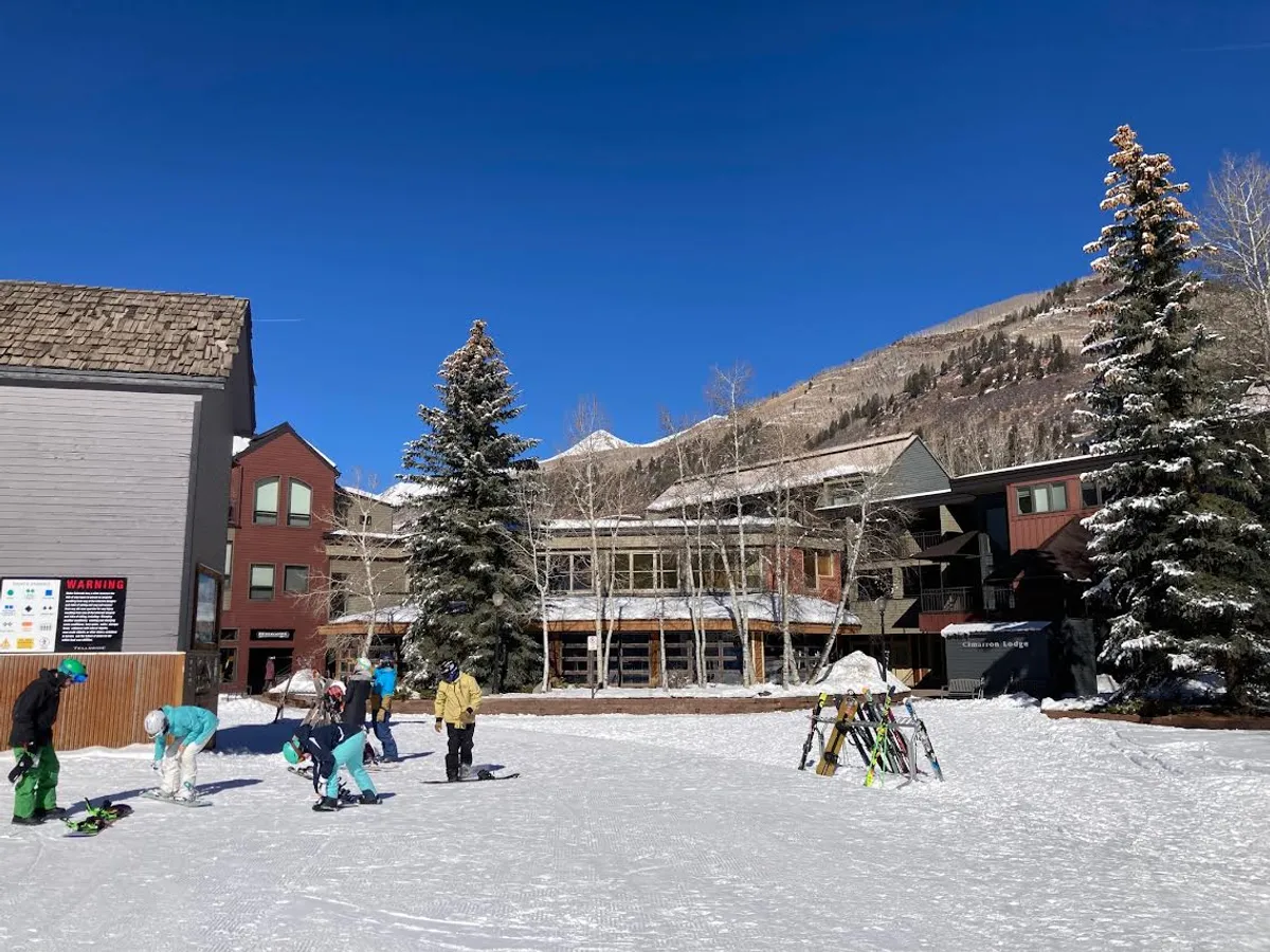 This image captures a dynamic scene of skiers preparing to head out on the snow-covered ground, with surrounding trees, a mountain backdrop, and buildings that evoke classic ski resort architecture. The atmosphere is lively and adventurous, perfectly encapsulating the excitement of ski-in/ski-out living.