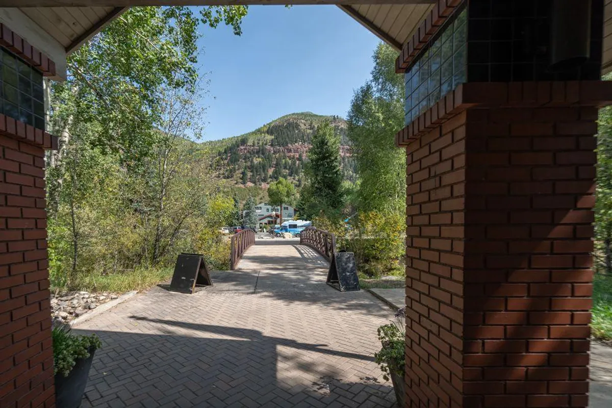 A picturesque arched entrance leads to a small bridge flanked by lush green trees, with mountains visible in the background. The path invites exploration, creating a sense of arrival and discovery amid tranquil surroundings.