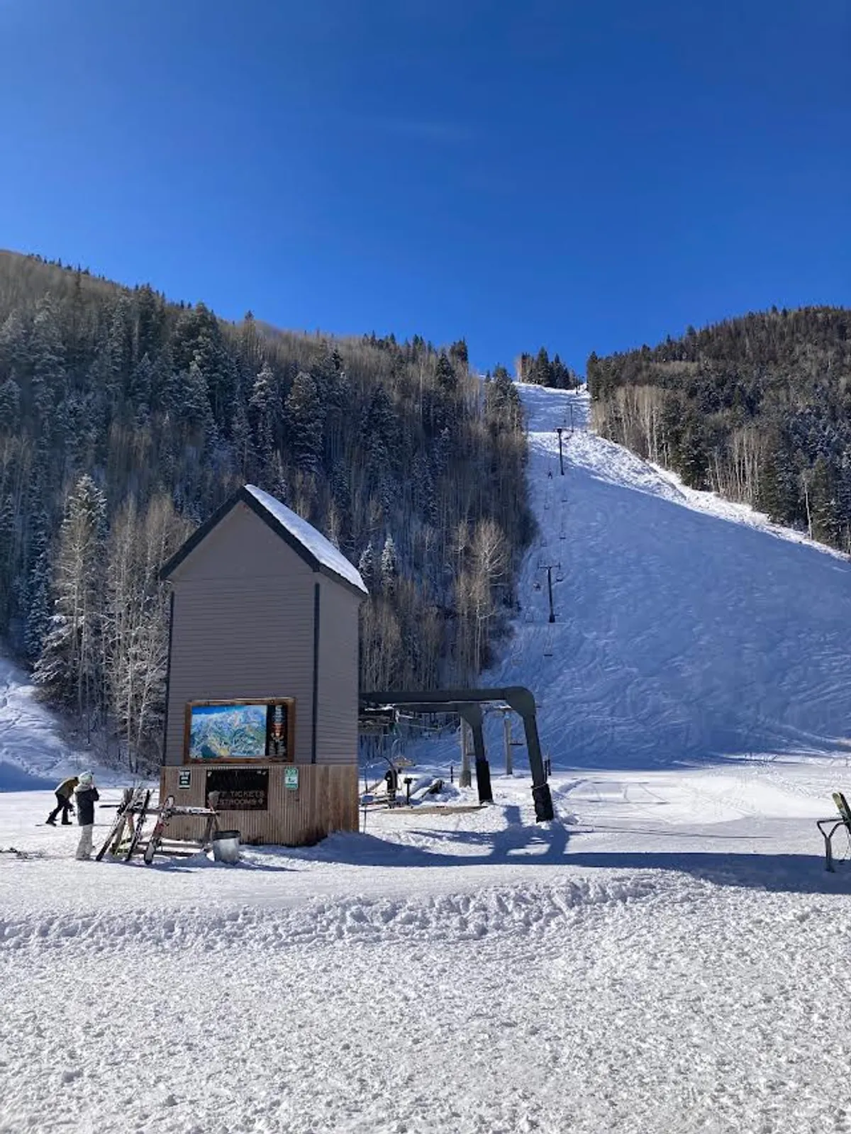 A dynamic exterior view featuring the ski lift and ticket booth, with people preparing to hit the snow-covered slopes. The image conveys the excitement of easy access to skiing, with the lift ticket window visible and a lively, adventurous mood.