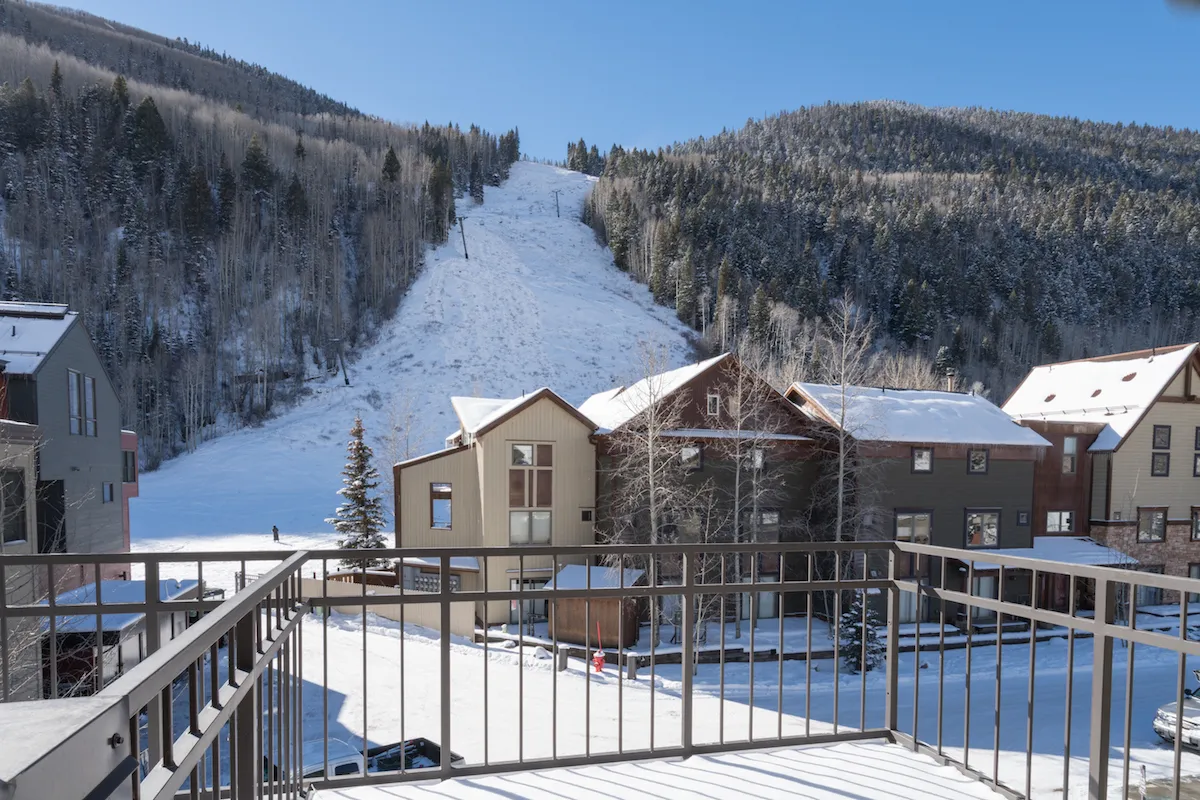 A tranquil winter scene featuring a snow-covered ski slope just beyond a balcony railing, framed by trees and buildings. The mountain view stretches toward the horizon, hinting at the adventure and peace found steps from the property.